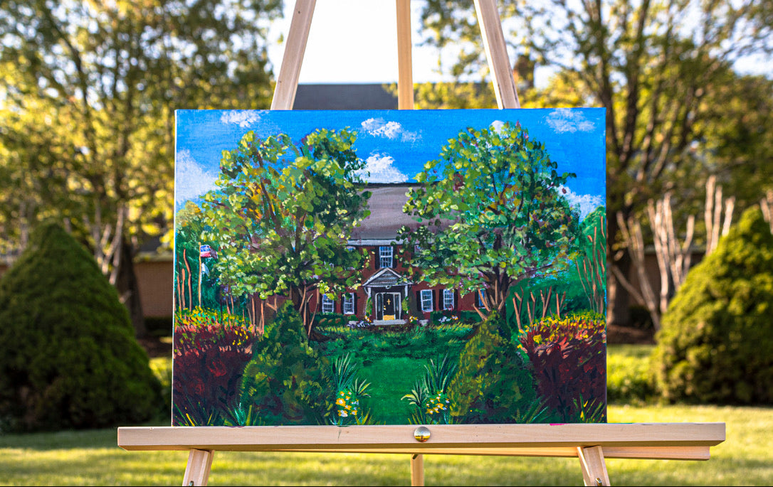 A colorful acrylic painting of a house surrounded by lush green trees and bushes under a bright blue sky, displayed on a wooden easel in an outdoor garden setting.