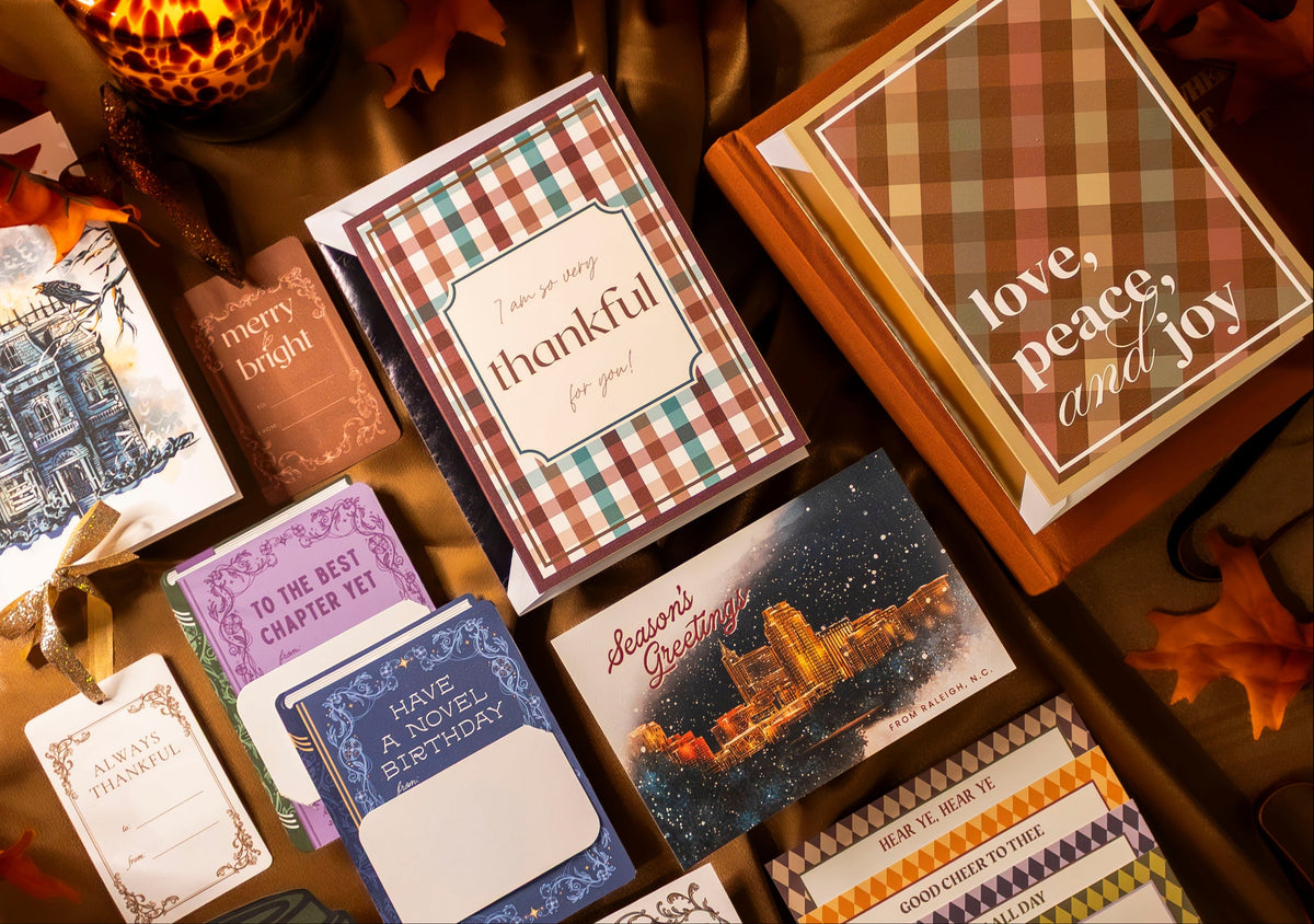 A styled flat-lay of multiple handmade greeting cards, stationery items, and a small wooden box arranged on a brown fabric surface with autumn leaves and a candle. The products include patterned cards, a card reading 'thankful,' a card with 'love, peace, and joy,' and assorted paper goods.