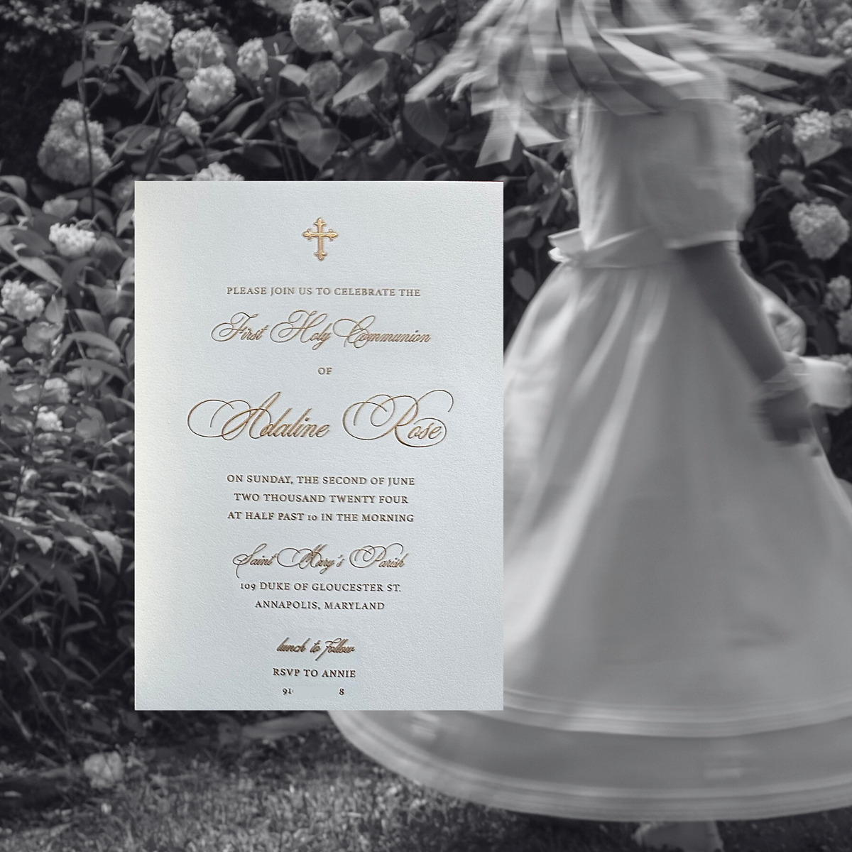 A first communion invitation is shown in front of a black-and-white photo of a child in a white dress standing in a flower garden. The invitation has a white background with elegant gold script and a small gold cross at the top.